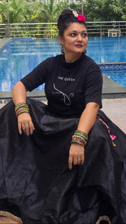 Woman in a black outfit with 'The Queen' text sitting by a pool with buildings and trees in the background.